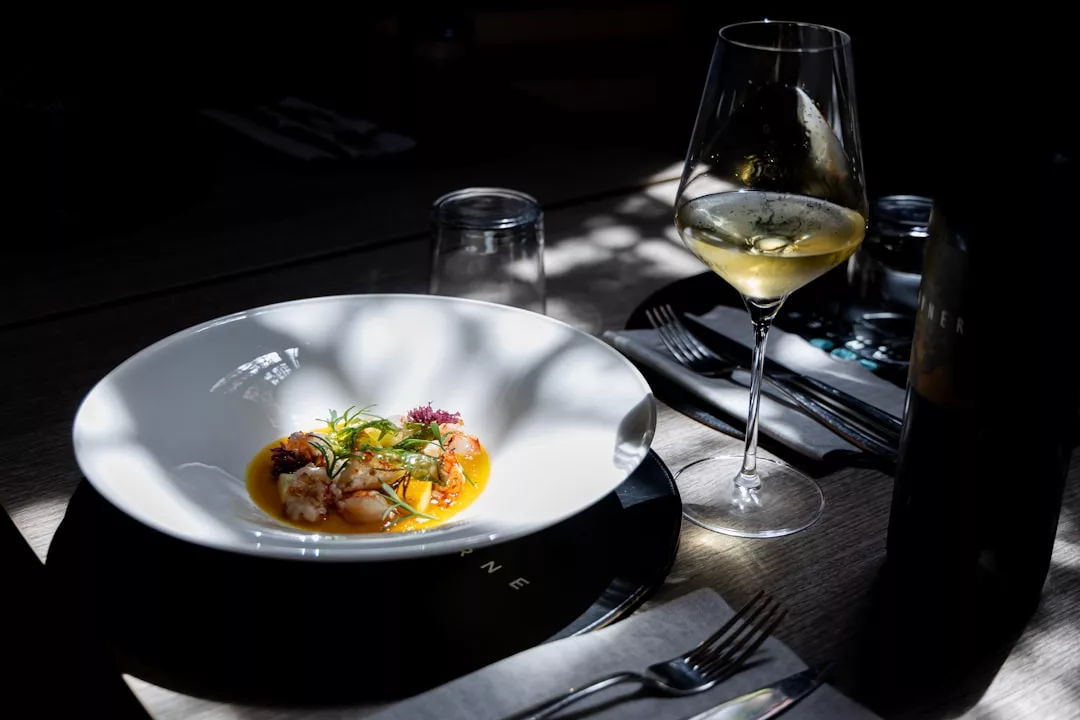 A white bowl with a seafood dish garnished with herbs sits on a table next to a glass of white wine, cutlery, and a water glass in dim, dappled lighting—an invitation to savor laid-back lifestyle elegance.