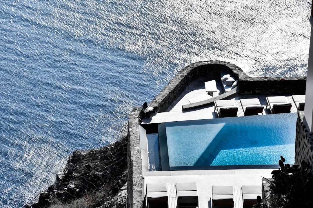 Experience coastal luxury with an infinity pool beside the sunlit ocean, surrounded by lounge chairs and a table—perfect for embracing the ultimate lifestyle in this stunning, panoramic setting.