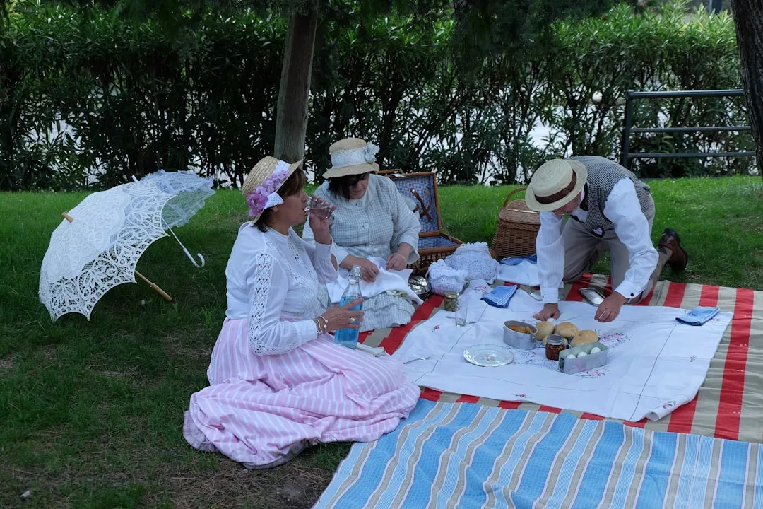 Three adults in vintage fashion enjoy a picnic on the grass, surrounded by food, drinks, and a lace parasol. A hedge and trees in the background complete this elegant lifestyle scene.