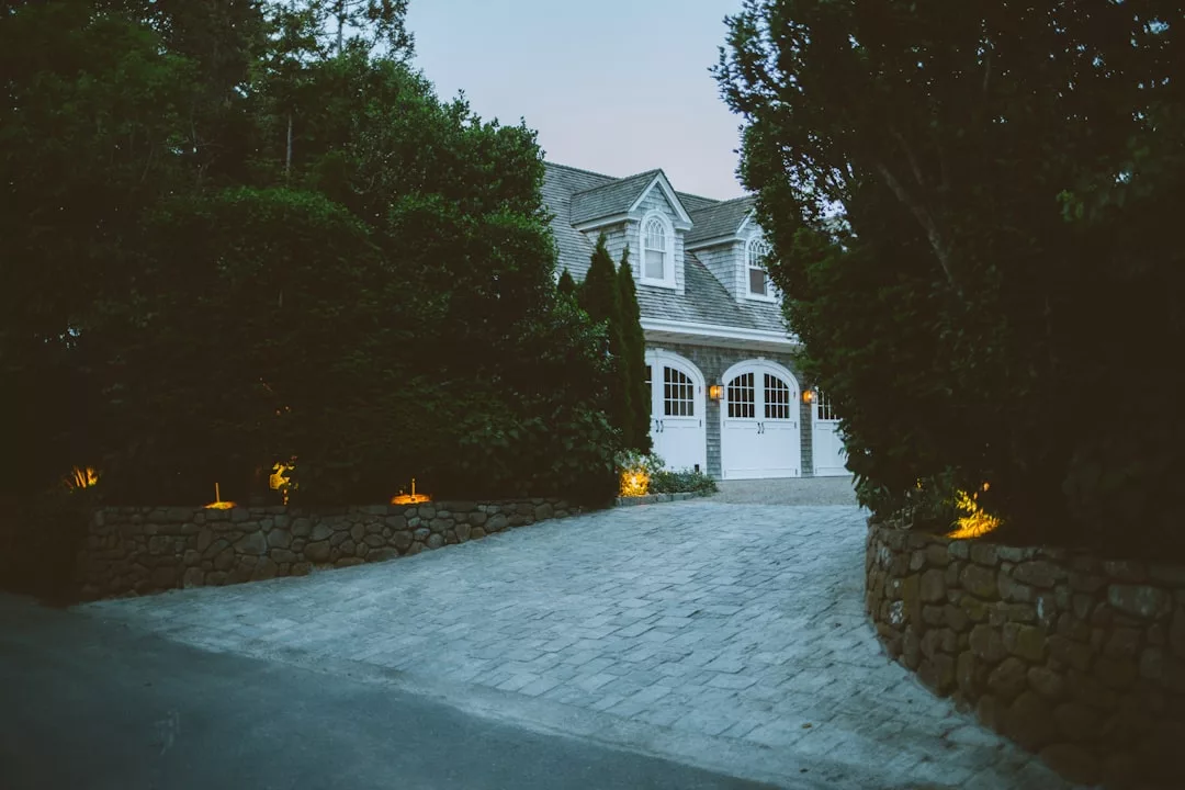 A paved driveway leads to a two-story luxury house with three white garage doors, surrounded by trees and illuminated garden lights at dusk.