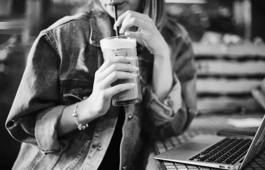 A person in a denim jacket enjoys a drink with a straw, embodying a relaxed lifestyle while sitting at a table beside an open laptop.