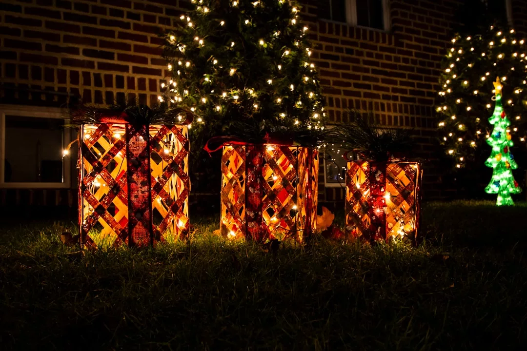 Four illuminated decorative gift boxes on grass in front of a lit Christmas tree exude a festive lifestyle, with a brick wall and a small glowing tree glowing warmly in the background at night.