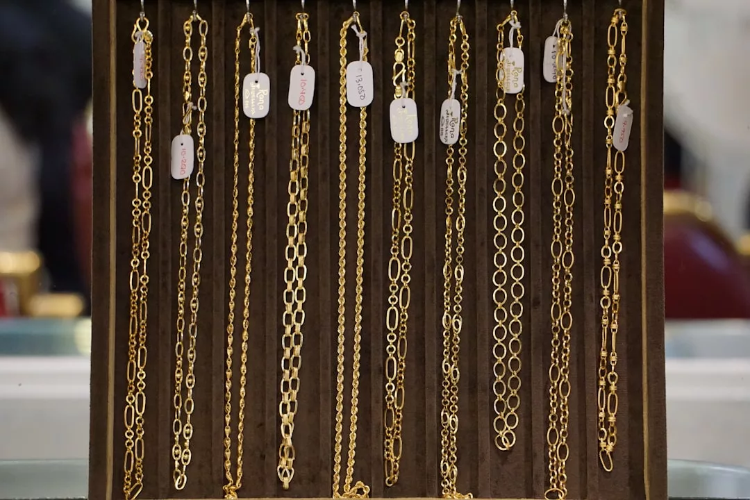 Several luxury gold chains of varying link styles and thicknesses hang vertically on display, each labeled with a white price tag, against a brown background.