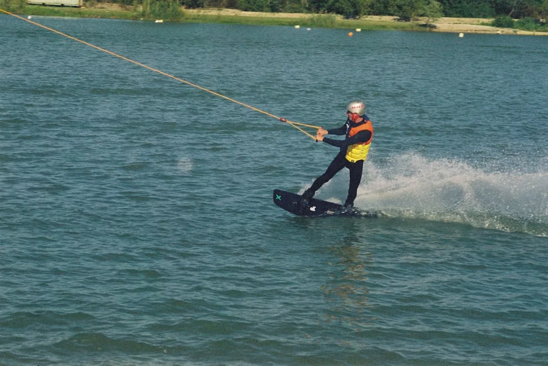 A person showcasing an adventurous lifestyle is wearing a helmet and life jacket while wakeboarding on a lake, holding onto a cable system.