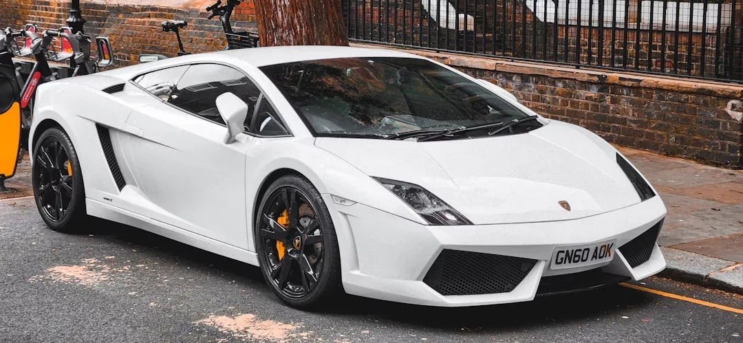 A white Lamborghini Gallardo, epitomizing luxury, is parked on a street beside a brick wall and metal fence, with bicycles visible in the background.