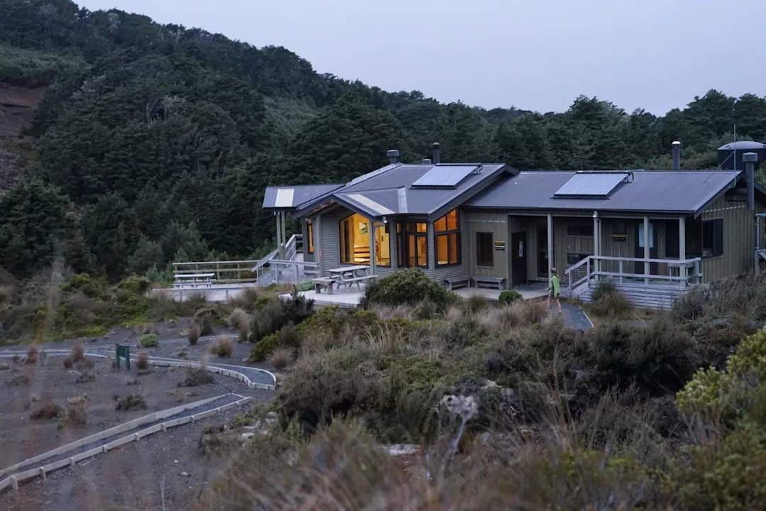 A modern luxury cabin with large windows and exterior lights sits surrounded by trees and shrubs, with a wooden deck and pathways visible in the foreground.