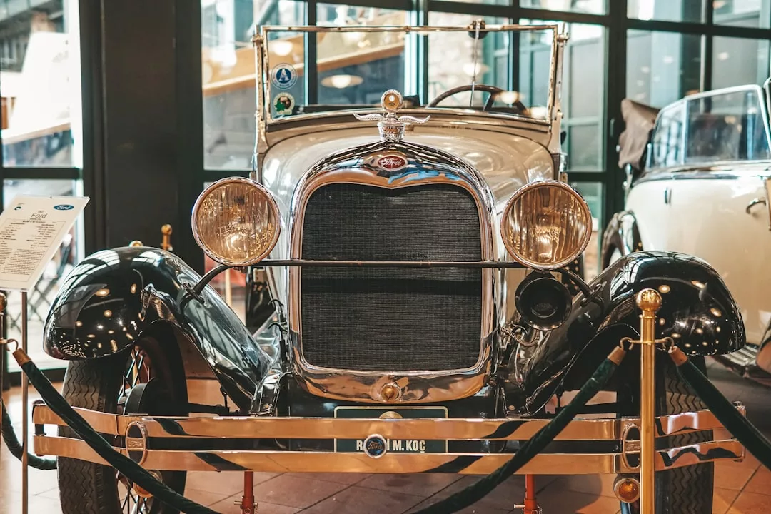 A vintage black and silver classic car with a front grille and round headlights is displayed indoors behind stanchions, exuding timeless fashion and Glamesia appeal.