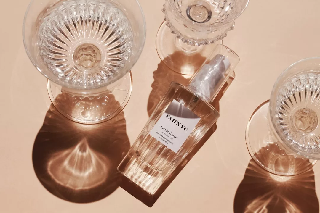 A clear glass bottle labeled "TAIWYC." sits on a peach-colored surface next to two empty textured glass cups, all casting distinct shadows—an understated nod to Glamesia style and modern luxury.