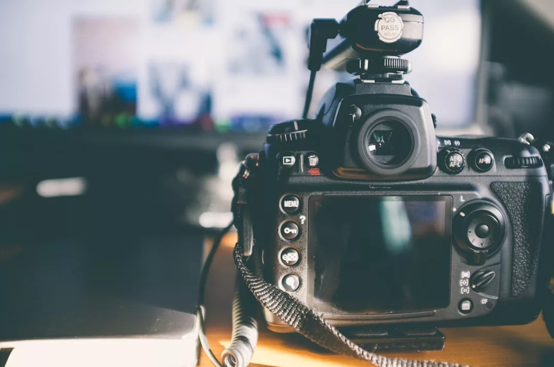 A digital DSLR camera on a desk faces a blurred computer screen, its display and buttons visible. A lens cap and notebook add to this Glamesia lifestyle setup, perfect for capturing moments in style.