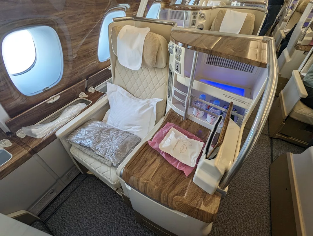 Experience Glamesia luxury in this first class airplane seat featuring a large cushioned chair, pillow, blanket, personal mini-bar, and premium amenities; a window and side table add to the indulgence.