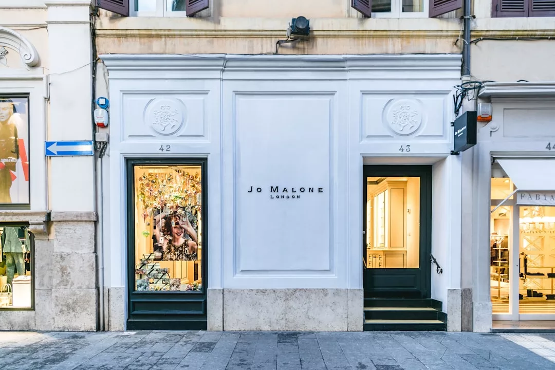 Storefront of Jo Malone London with white facade, flanked by large display windows and dark green doors, offering a touch of fashion and lifestyle luxury at street numbers 42 and 43.