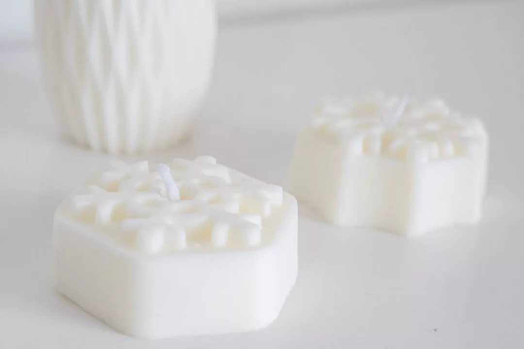 Two white, hexagonal wax melts with snowflake patterns rest on a white surface, exuding a sense of luxury, while a textured white container sits elegantly in the background.