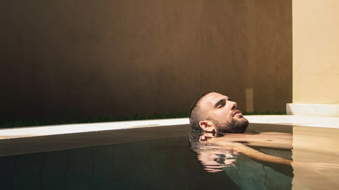 A man enjoys a Glamesia lifestyle, relaxing in a swimming pool with his head resting on the edge, eyes closed as sunlight casts soft shadows on the wall behind him.