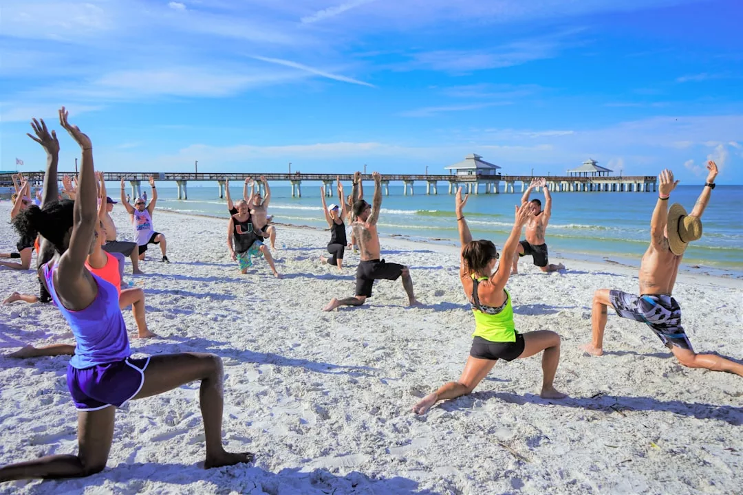 A group of people practice yoga on a sandy beach near the ocean, embracing a Laxary lifestyle with Glamesia-inspired athleisure, as a pier and blue sky complete the serene backdrop.