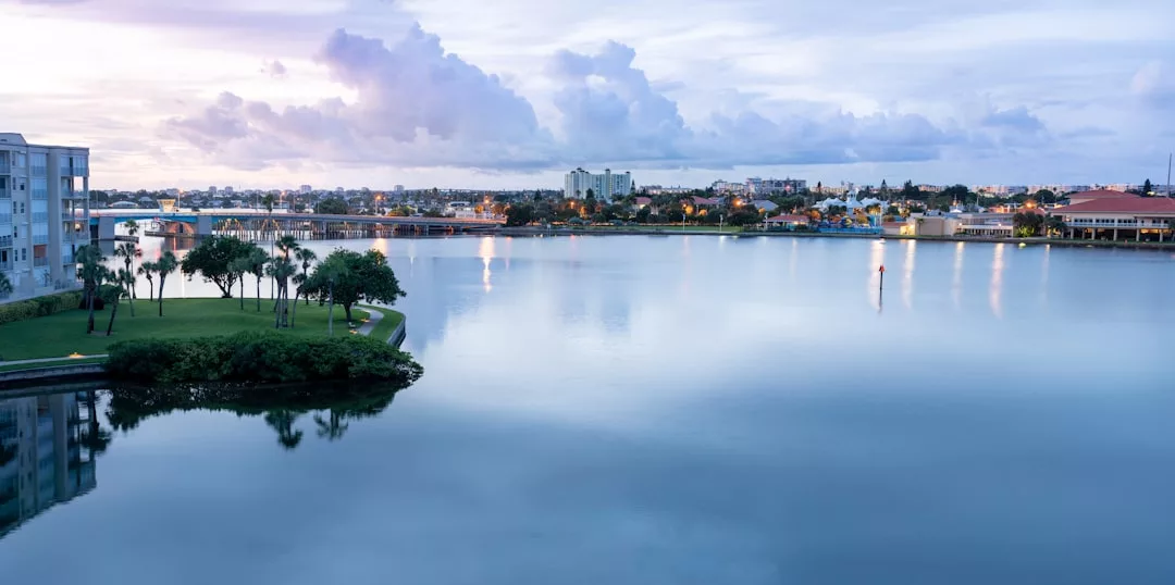 A calm waterfront scene at dusk with luxury buildings, trees, and lights reflected in the water under a cloudy sky.