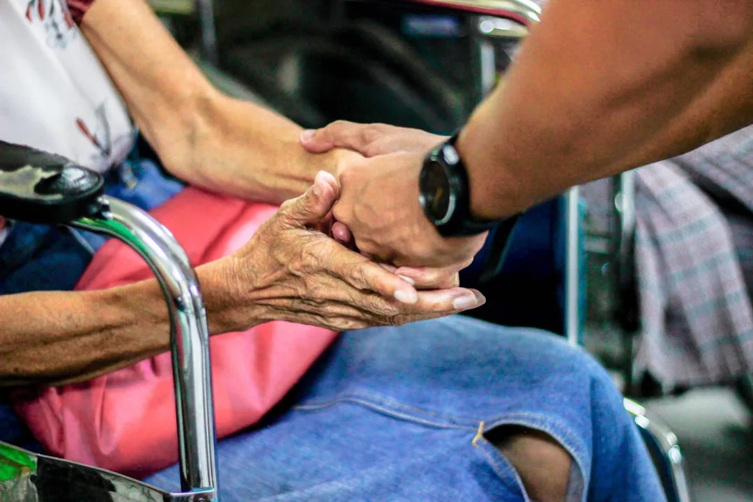 A person showcasing lifestyle fashion wears a watch while gently holding the hands of an elderly individual sitting in a wheelchair with a pink bag on their lap.
