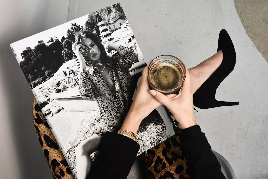 Person seated with legs crossed, holding a drink in one hand and a black-and-white fashion magazine featuring a woman in leopard print in the other; showcasing luxury lifestyle with leopard print pants and sleek black high heels.
