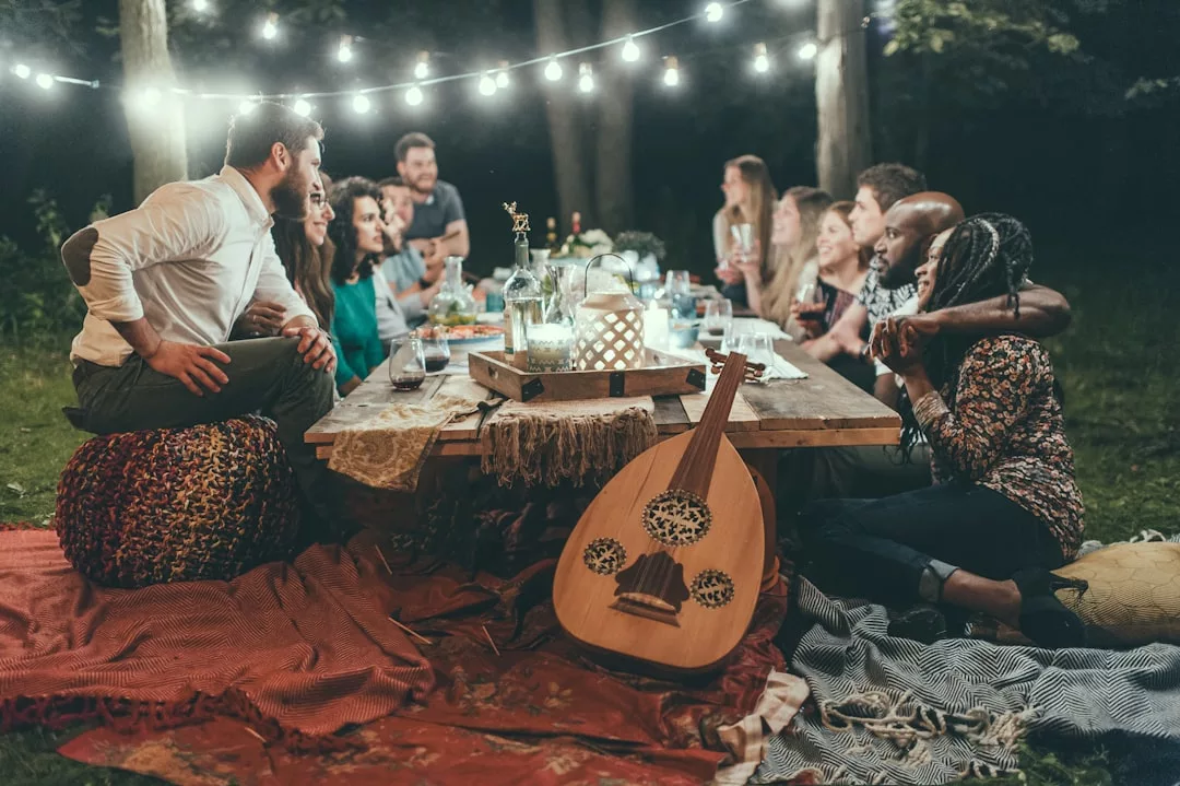 A group of people sit around a low outdoor table at night, sharing food and drinks under string lights, with a hint of Glamesia luxury in their setting and a lute resting on a blanket in the foreground.