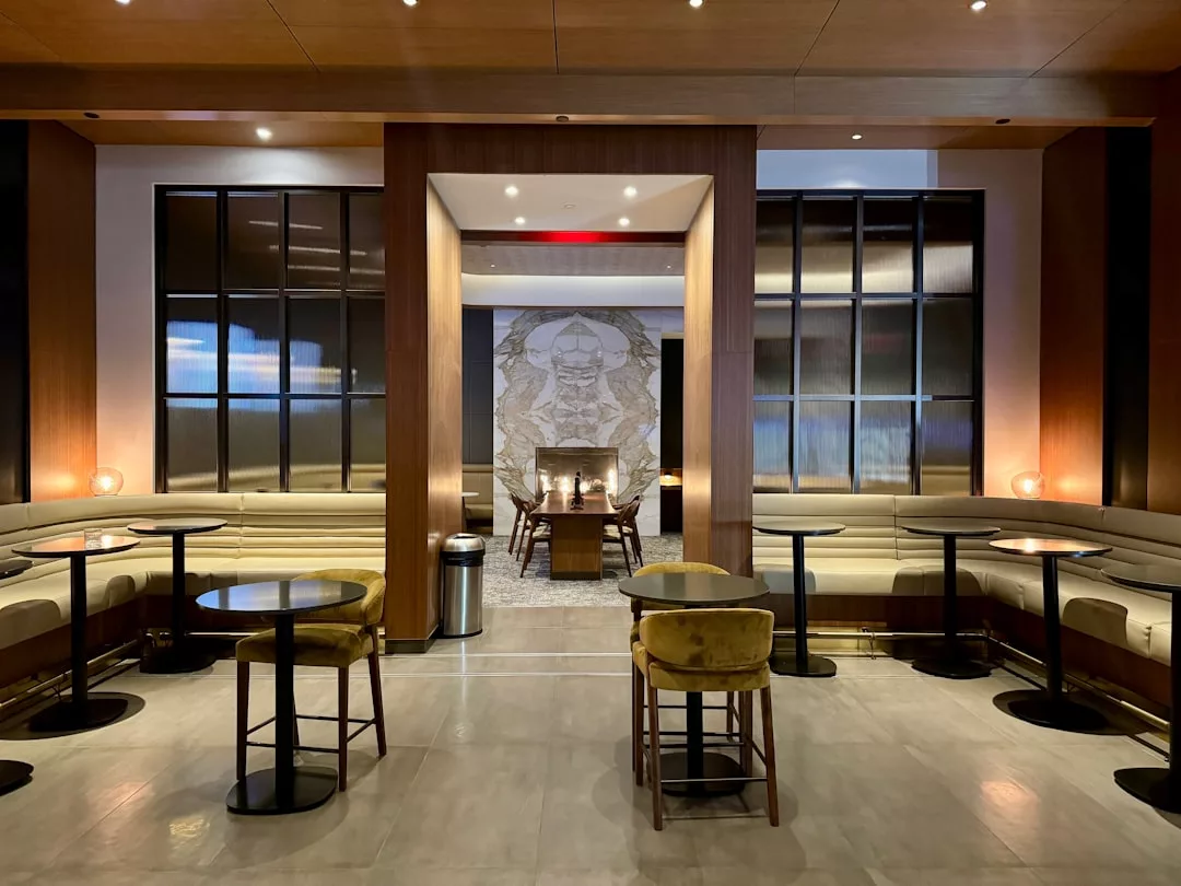 Modern lounge area with beige banquette seating, round tables, velvet chairs, and a wall mural in the background under warm lighting—an inviting space that blends luxury and lifestyle for a fashionable social setting.