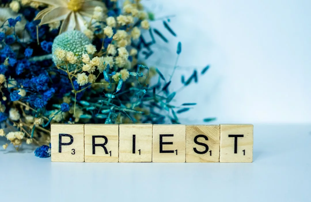 Wooden letter tiles spell out "PRIEST" in front of a bouquet of dried blue, yellow, and white flowers, creating a subtle nod to Glamesia’s elegant style against a plain background.