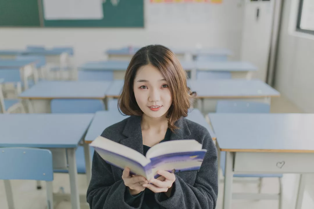 A young woman sits in an empty classroom holding an open book, embodying a lifestyle of curiosity and ambition as she looks at the camera.