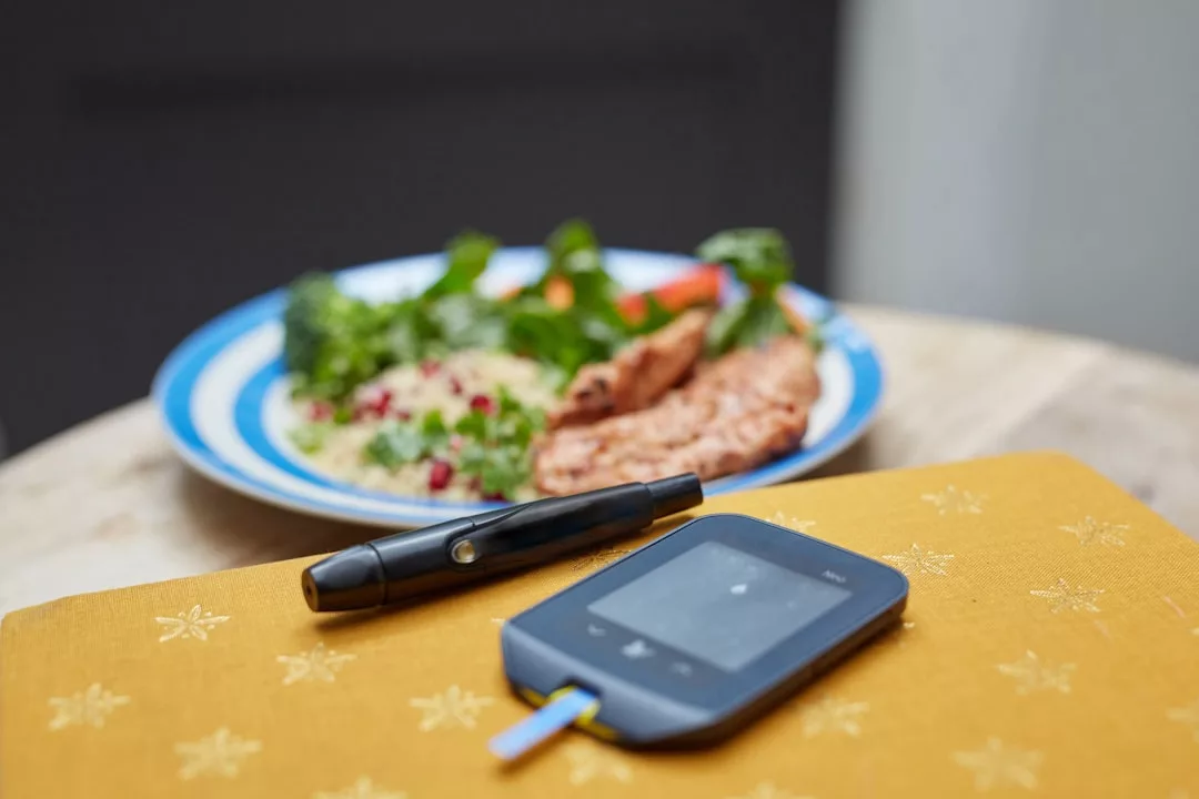 A blood glucose meter and lancing device sit on a table—an essential part of a healthy lifestyle—with a plate of vegetables and protein blurred in the background.