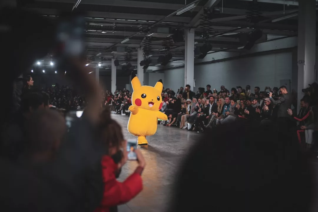 A person in a Pikachu costume walks down a fashion runway at an indoor event, with a seated audience watching and some people taking photos.