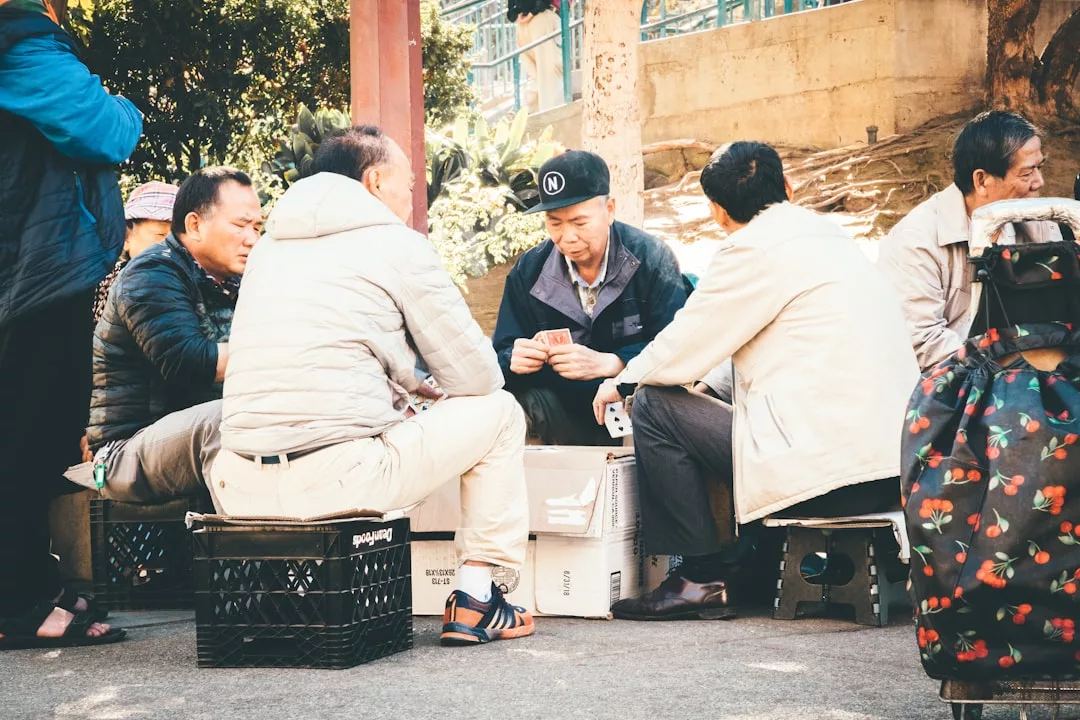 A group of men sits on crates and boxes outdoors, playing cards around a makeshift table, while another person with a rolling bag watches the scene unfold in this moment far removed from luxury.