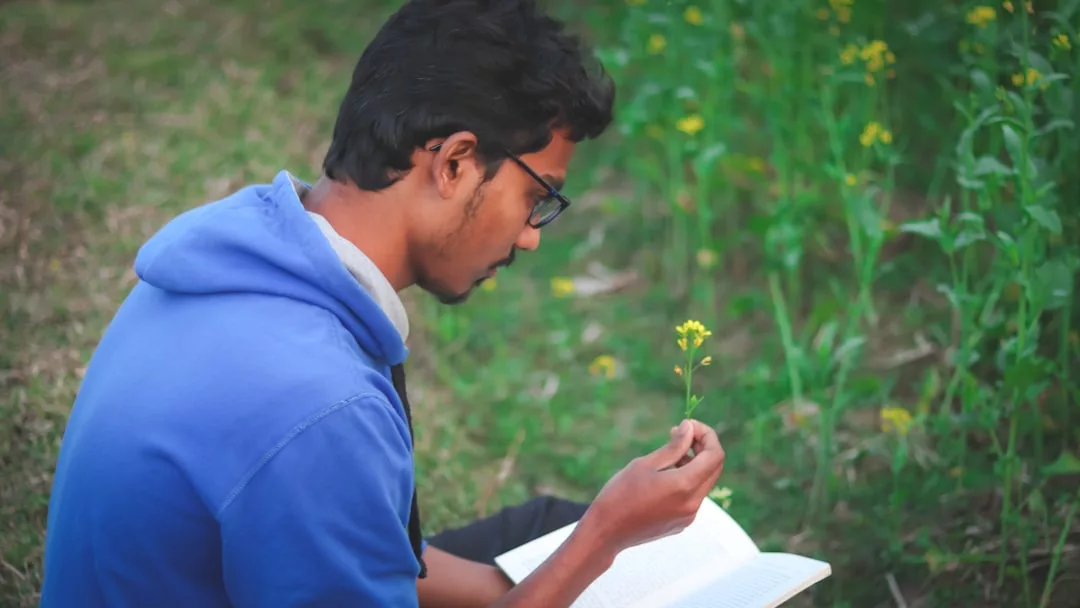 A man with glasses and a blue hoodie sits outdoors, holding a flower in one hand and an open book in the other, showcasing relaxed fashion amid lush green plants.