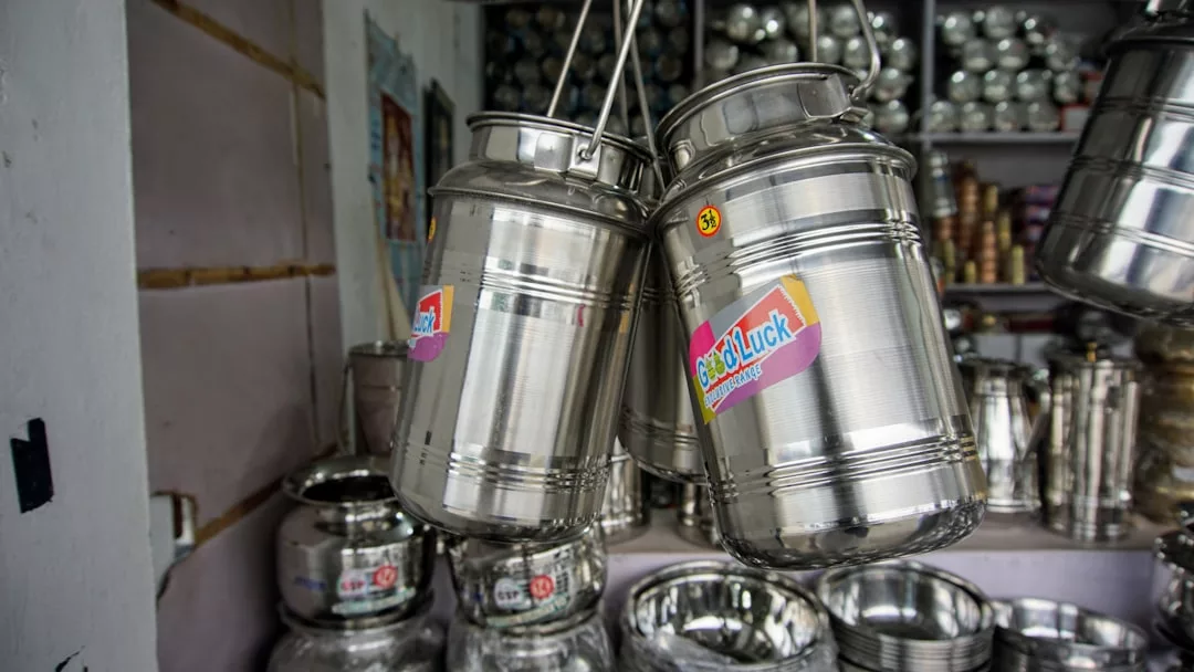 Two shiny stainless steel milk containers with "Good Luck" labels hang in a shop, surrounded by other metal kitchenware and utensils, adding a touch of lifestyle elegance to the scene.