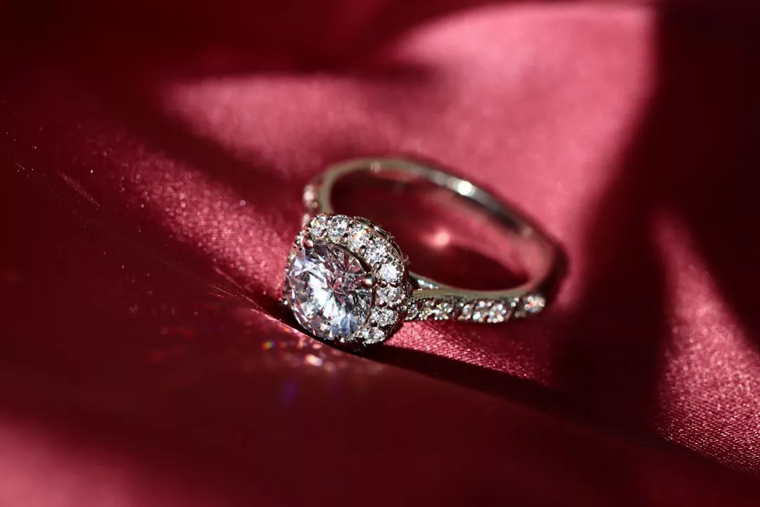 A luxury diamond engagement ring with a round center stone and a halo of smaller diamonds rests on red satin fabric, radiating elegance.