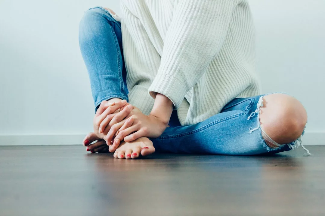 Person sitting on the floor with legs crossed, wearing a white sweater and ripped blue jeans, holding their bare foot with both hands—a relaxed lifestyle look blending comfort and fashion.