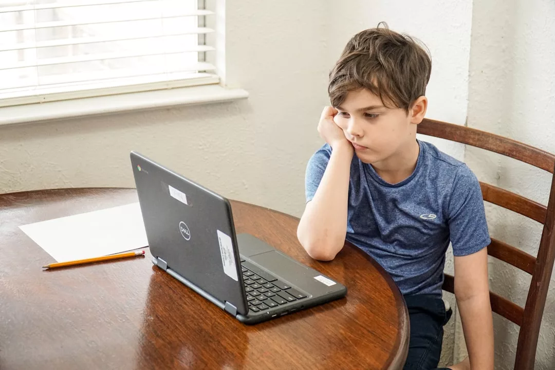 A boy in a blue shirt sits at a table, looking at a laptop with his head resting on his hand; beside him, a pencil and paper add a touch of quiet focus to this Glamesia moment.