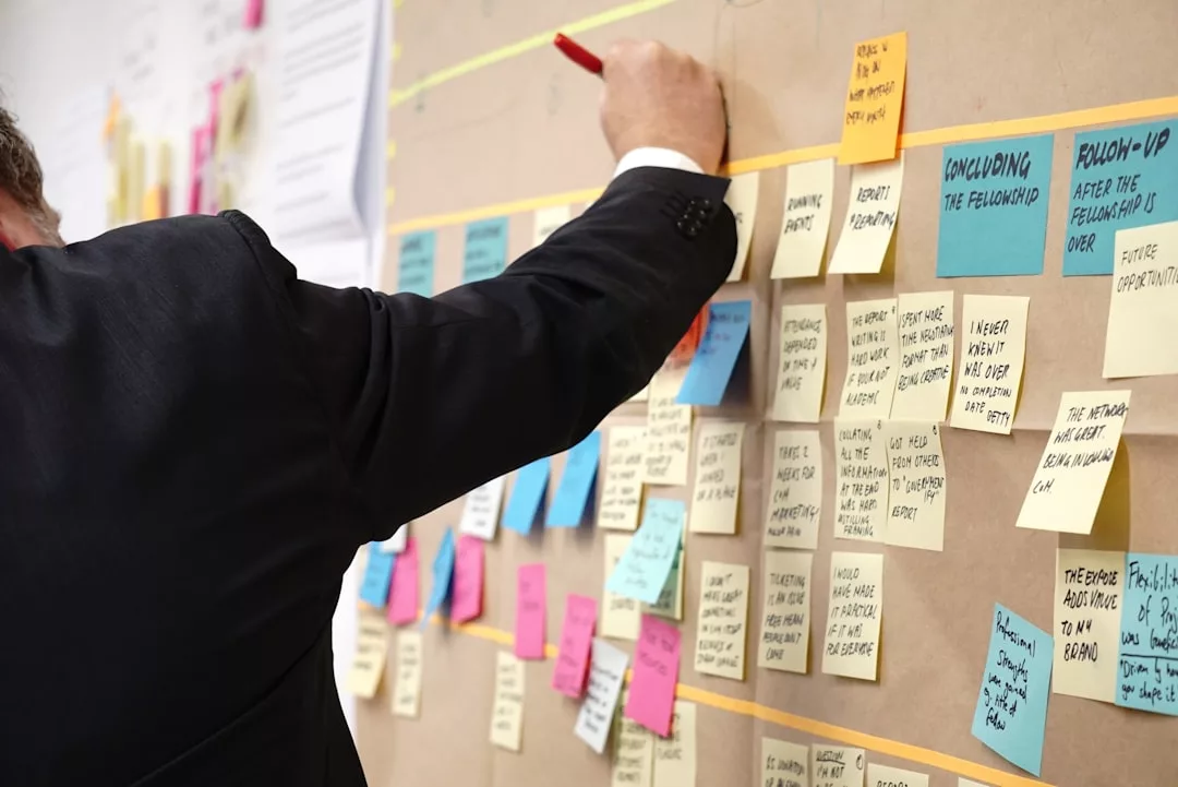 A person in a suit writes on a sticky note on a board covered with colorful, handwritten notes, reflecting an organized luxury lifestyle.