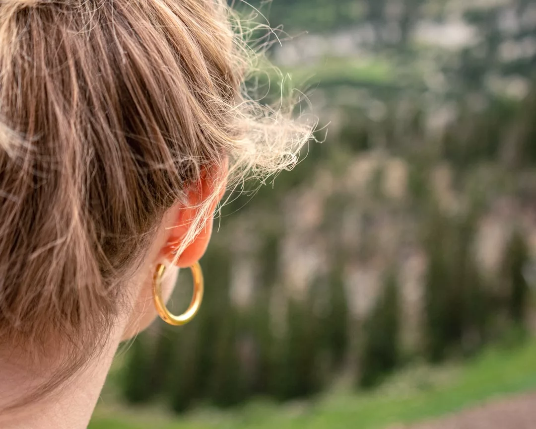 Close-up of the back of a person's head with light brown hair in a ponytail, wearing a gold hoop earring—a touch of fashion—while gazing out at a blurred forested landscape.