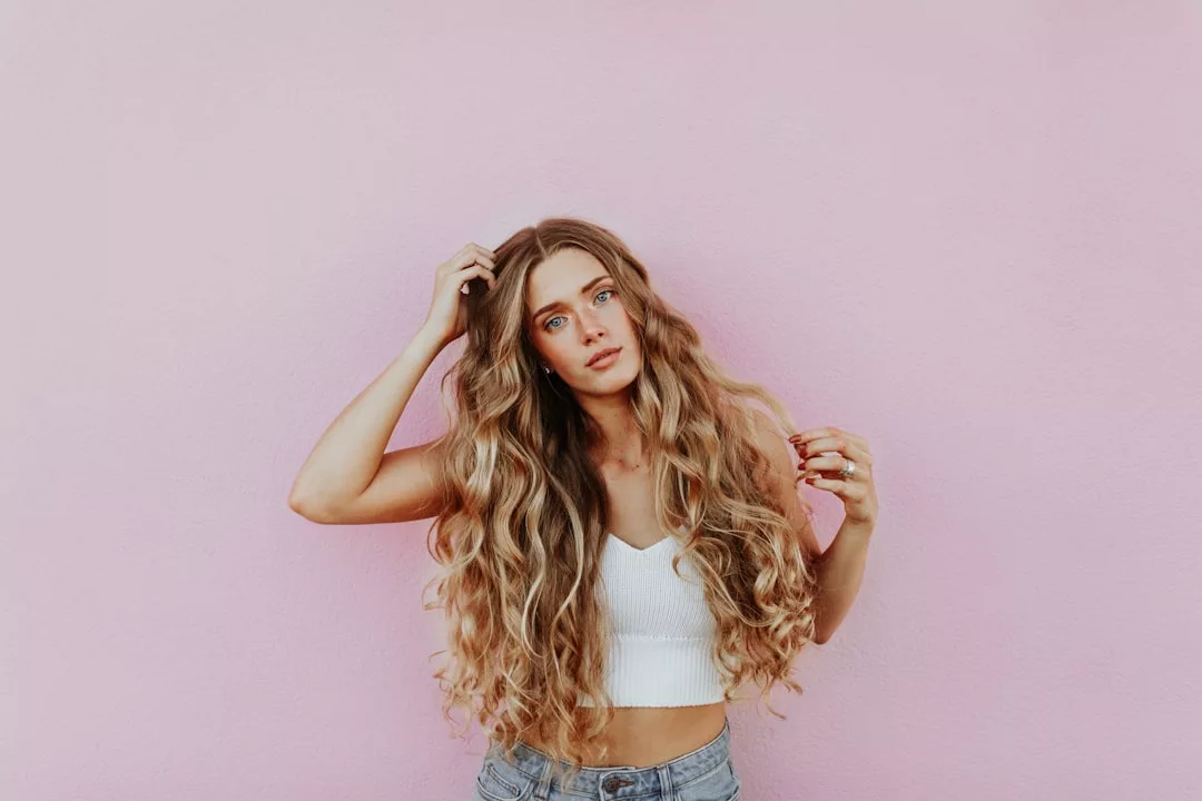 A person with long, wavy blonde hair stands against a light pink wall, showcasing effortless fashion in a white cropped tank top and blue jeans.
