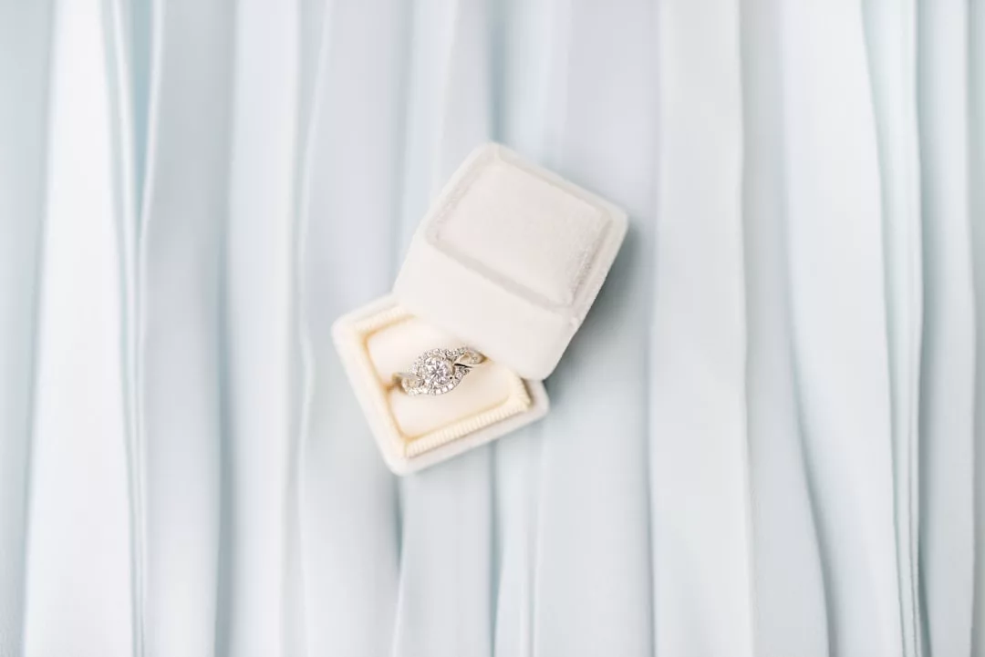 A diamond engagement ring in a white velvet box is displayed against a light blue, pleated fabric background, capturing a sense of fashion and refined lifestyle.
