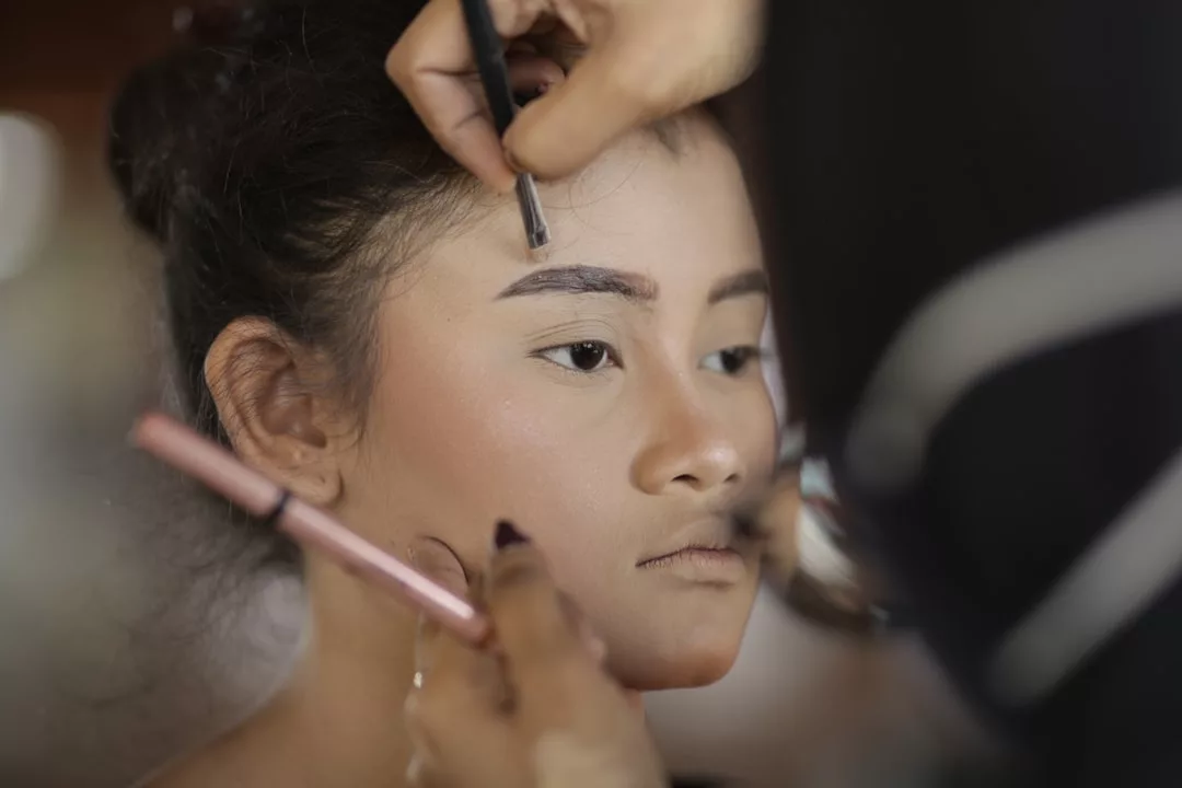 A person enjoys a luxury makeup session as one hand shapes their eyebrow with a pencil while another applies product on the cheek with a brush—a true Glamesia lifestyle moment.