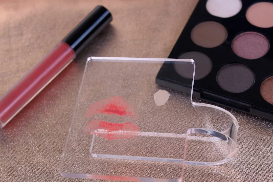A clear acrylic coaster with a lipstick kiss mark rests on a shimmery surface beside a lip gloss tube and eyeshadow palette, creating the perfect Glamesia lifestyle vibe.