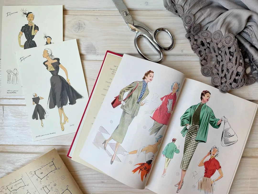 An open Glamesia fashion sketchbook with vintage clothing illustrations, fabric, scissors, sewing patterns, and two luxury fashion postcards on a wooden table.