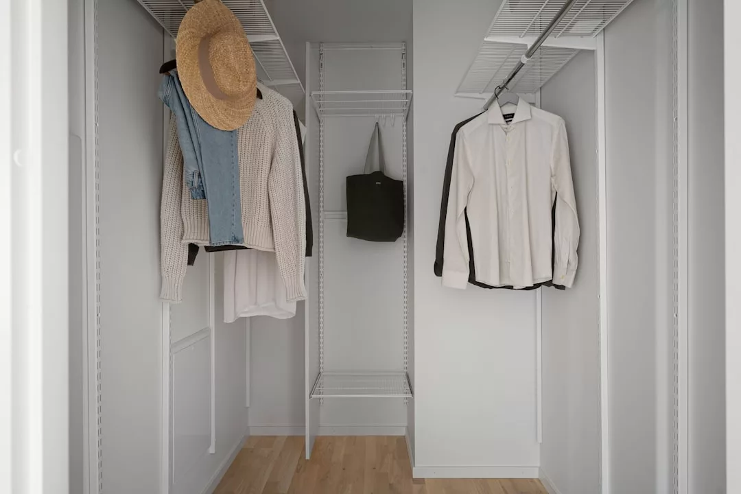 A luxury walk-in closet with hanging clothes, a straw hat, a green tote bag, and empty wire shelves against pristine white walls and a sleek wood floor.