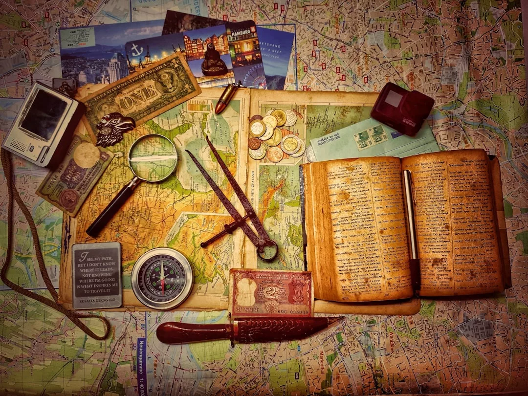 A vintage travel-themed flat lay with an old map, open journal, compass, magnifying glass, foreign currency, coins, camera, and postcards arranged on a map background evokes timeless luxury and adventurous fashion.