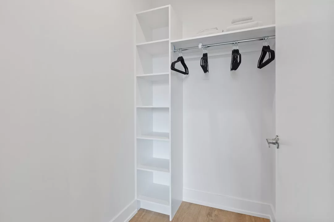 Empty white closet with open shelving, a hanging rod holding several black hangers, two folded towels on the top shelf, and a light wooden floor—perfect for organizing your fashion essentials in a modern lifestyle setting.