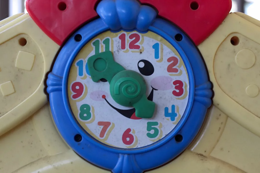 A colorful toy clock with a smiling face features green movable hour and minute hands pointing at 10 and 12, set on a yellow and blue plastic frame—perfect for adding fun style to any lifestyle.