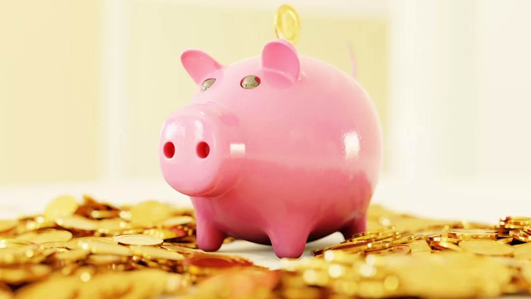 A pink piggy bank with a coin being inserted on top, surrounded by scattered gold coins on a light surface, adds a touch of Glamesia style to your savings lifestyle.