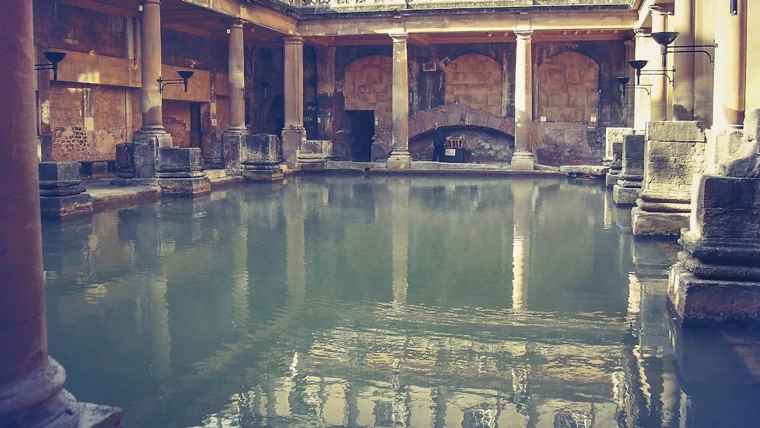 Ancient Roman-style bath with stone columns and walls surrounding a large rectangular pool of water indoors, creating a luxurious lifestyle experience.