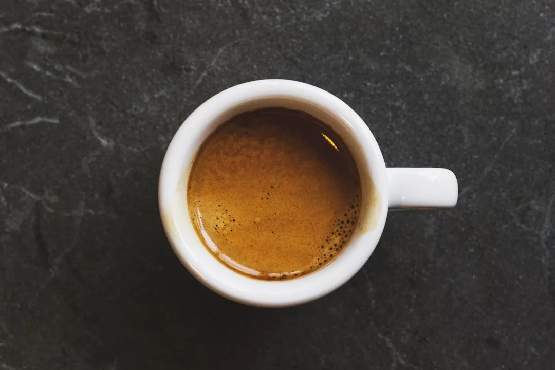 A white ceramic cup filled with espresso sits on a dark stone surface, viewed from above—a simple moment reflecting contemporary lifestyle and understated fashion.