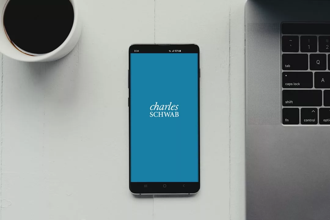 A smartphone displaying the Charles Schwab app screen sits on a white desk next to a cup of coffee and a partially visible laptop, creating a modern lifestyle scene.