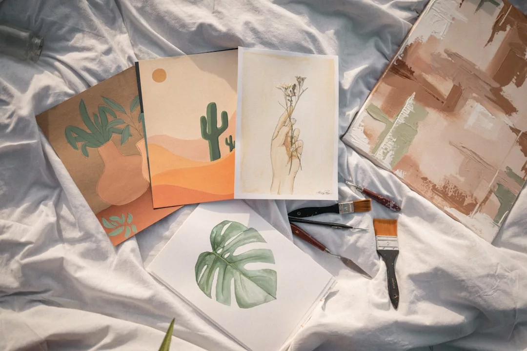 Several art prints featuring plants and abstract designs are displayed on a white sheet alongside three paintbrushes, adding a touch of luxury to your lifestyle décor.