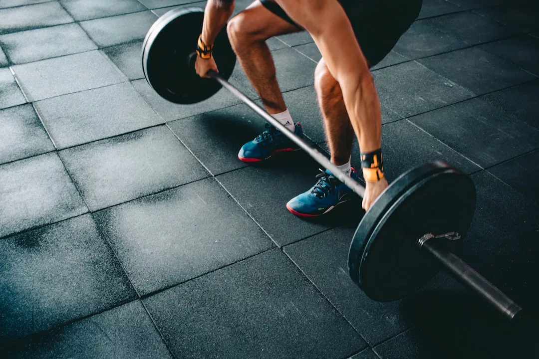A person sporting fashionable blue shoes prepares to lift a barbell with black weight plates on a gym floor with black tiles.